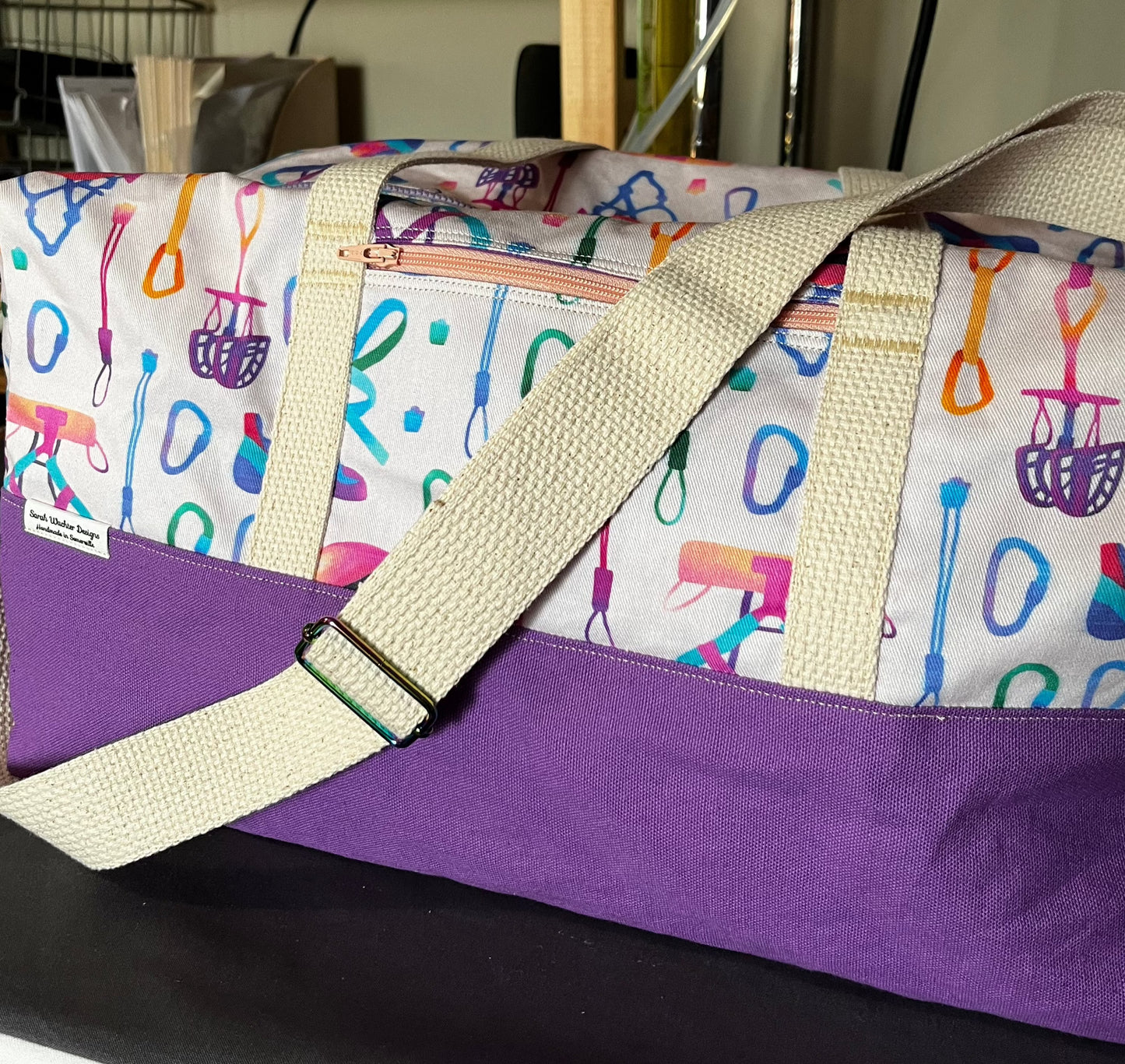 A duffel bag with a lilac base and natural colored shoulder and tote straps with rainbow hardware. The top portion of the bag is printed with a brightly rainbow-colored pattern of climbing gear such as carabiners, nuts, harnesses, climbing shoes, etc on a pale peach background.