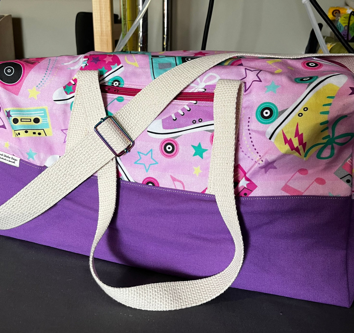 A duffel bag with a lilac base and natural-colored shoulder and tote straps.  The upper portion of the bag has scattered brightly colored roller skates, cassette tapes, and retro stereos on a pale pink background.