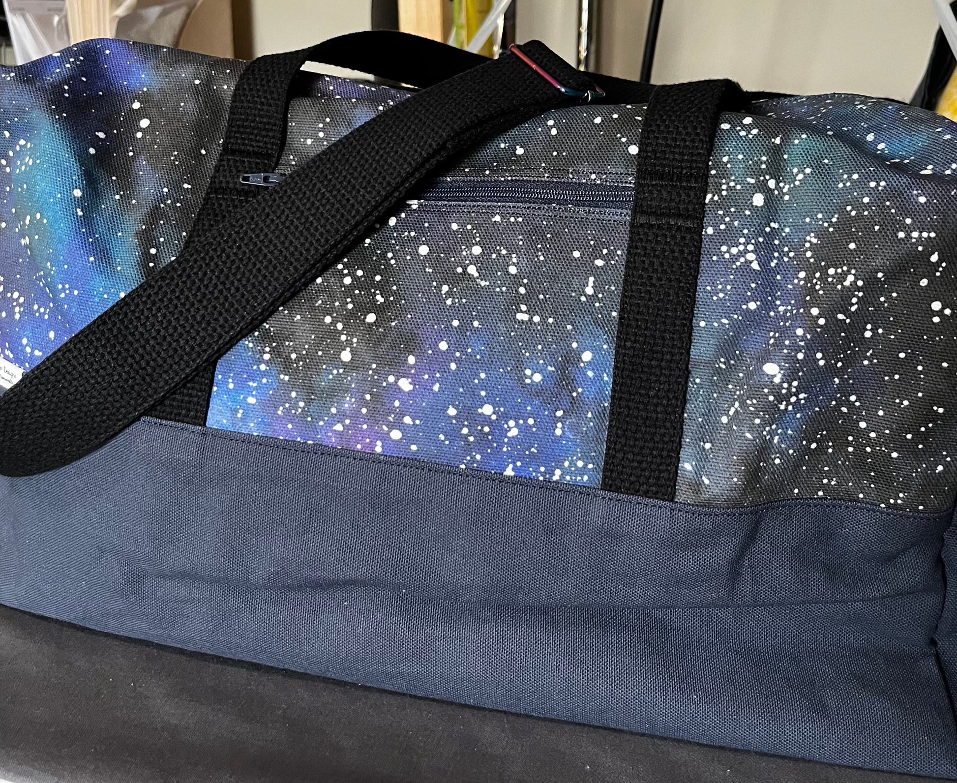 A duffel bag with a navy blue base and black shoulder and tote straps with rainbow hardware.  The top portion of the bag is printed with a starry sky in a nebula-type effect on a mostly black background.