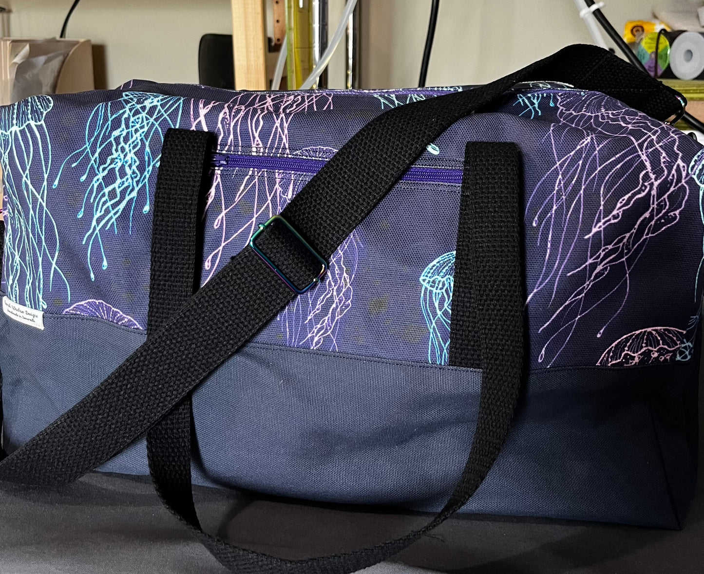 A duffel bag with a navy blue base and black shoulder and tote straps with rainbow hardware.  The top portion of the bag has a pattern of brightly colored blue and purple jellyfish on a deep blue-black background.